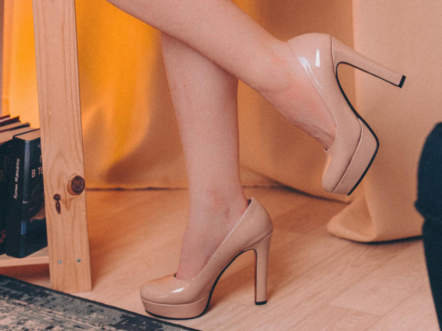 Decadance's legs in glossy nude high heels, crossed at the ankle, showing off her shiny, beige pumps with a subtle heel. Her skin is fair, and the wooden floor is visible.