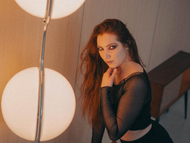 Decadance with long brown hair, wearing a sheer black top, touches her chin, has red eyes, and a serious expression, standing near two glowing spherical lights.