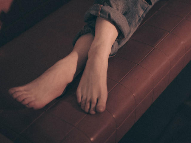 I piedi nudi di Decadance sono incrociati su una panca in pelle marrone, con i jeans grigi arrotolati che spuntano alle caviglie. Una luce soffusa e calda mette in risalto la loro pelle chiara..