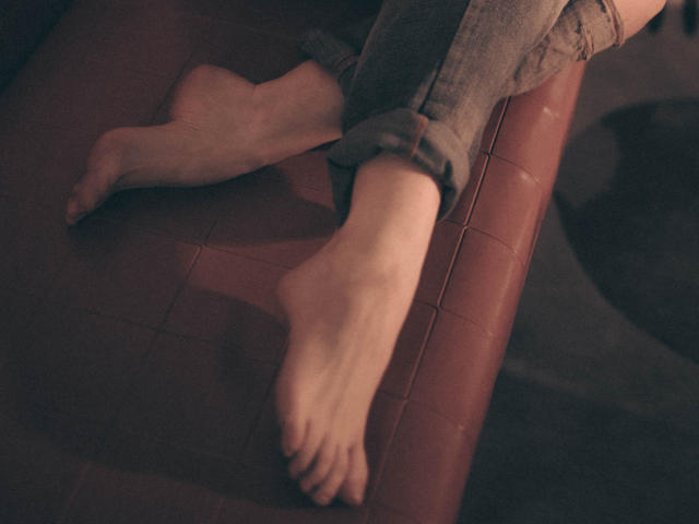 I piedi nudi di Decadance erano incrociati su una superficie di pelle rossa, con le gambe infilate in jeans grigi leggermente arrotolati, che trasmettevano un'aria rilassata e informale. Una luce soffusa e calda accentua l'atmosfera accogliente..