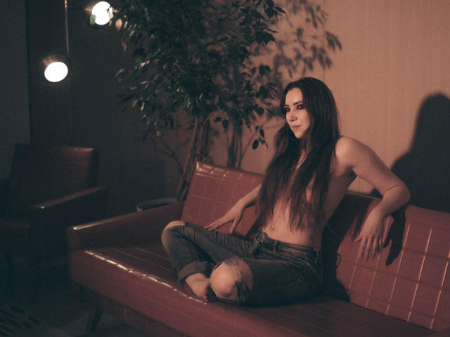 Decadance with long dark hair, topless, showing small tits, sits on a red couch in dim light, wearing ripped jeans, arms resting on the back, legs crossed, looking forward.