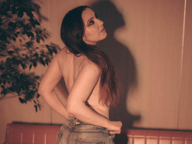Decadance with long brown hair, topless, looking over shoulder, hands in jeans, shadow on wall, puffy lips, slight smile, soft light, bare back, subtle makeup.
