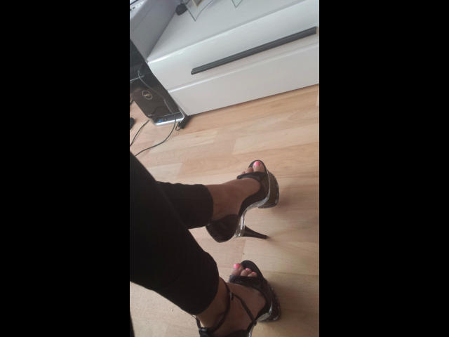 Le gambe nere di Minnieme, vestita di scuro e con i tacchi alti, mostrano le unghie dei piedi rosa, mentre è in piedi su un pavimento di legno. Sullo sfondo, si vedono un termosifone bianco e un cavo di alimentazione. L'immagine ha un'angolazione casuale, leggermente dal basso..