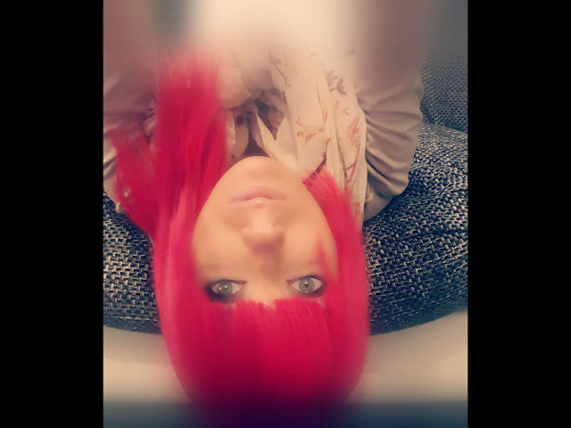 Minnieme-hot with bright red hair, upside down, looking at the camera with blue eyes. She's wearing a floral shirt, sitting on a blue, textured chair. Boobless, no visible pussy.