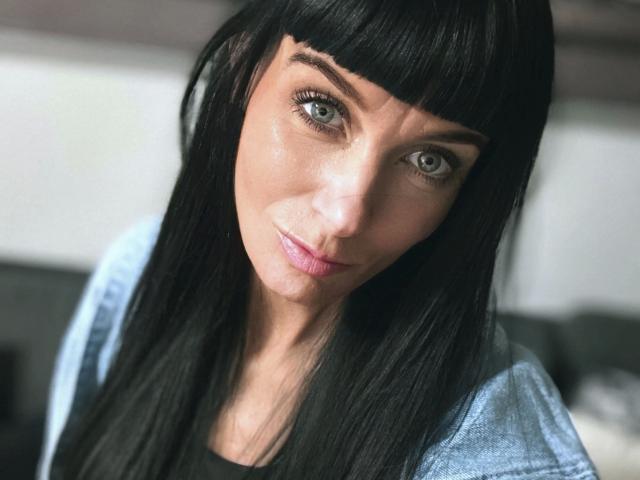 Minnieme, con i capelli neri lisci, gli occhi azzurri e le labbra rosa, indossa una camicia azzurra chiara, ha un sorriso delicato e la testa leggermente inclinata, illuminata da una luce soffusa e naturale..