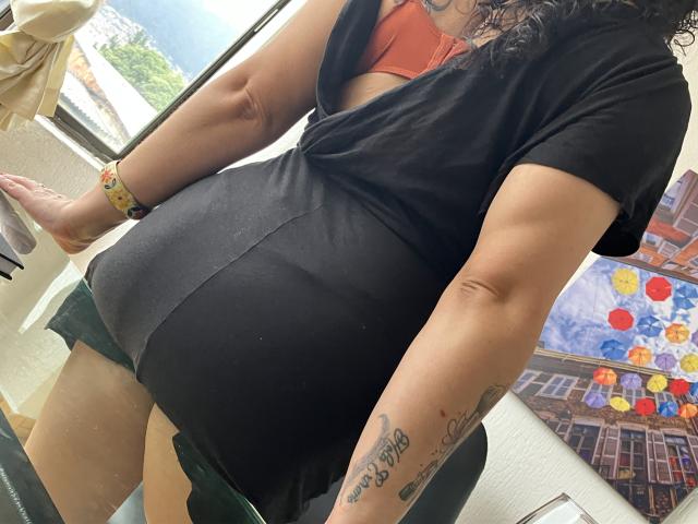 Tamarajoones, avec ses cheveux noirs bouclés, porte une robe noire et un soutien-gorge rouge, exposant ses fesses volumineuses et ses tatouages sur son bras droit. Elle est appuyée contre une fenêtre..