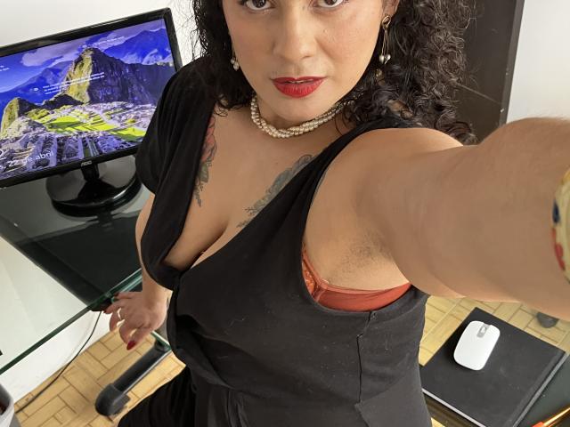 Tamarajoones con capelli neri ricci, rossetto rosso, vestito nero, collana di perle, che mostra i peli delle ascelle, rivela un reggiseno arancione e ha dei tatuaggi, sta scattando un selfie, c'è una TV sullo sfondo e un computer sulla scrivania..