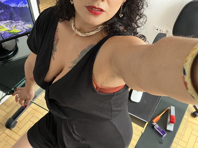 Tamarajoones mit lockigem, schwarzem Haar, rotem Lippenstift, schwarzem Kleid, einer Perlenkette, die ihre Brüste in einem roten BH zeigt, macht ein Selfie. Im Hintergrund sind ein Holzboden, ein Fernseher und ein Stuhl zu sehen..