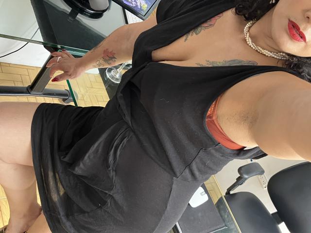 Tamarajoones indossa un abito nero con un profondo scollo, che mostra i suoi seni in un reggiseno rosso, ha tatuaggi sul braccio, una collana di perle e rossetto rosso, ed è seduta a una scrivania..