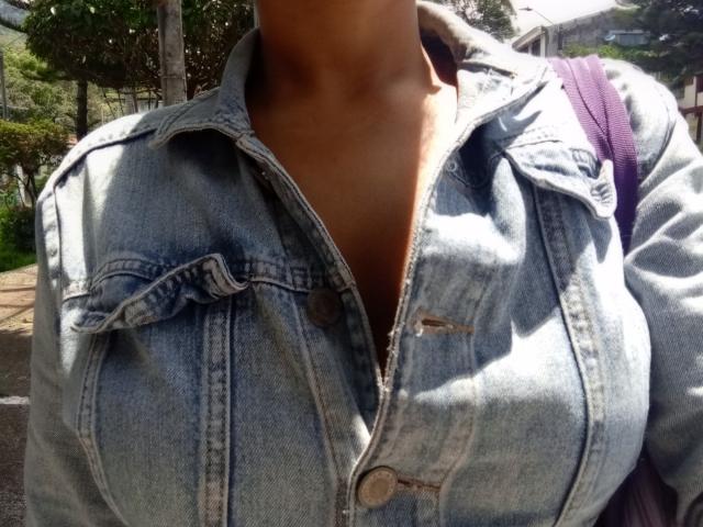 MeganWilliams' large tits are covered by a slightly unbuttoned, light blue denim jacket with brown buttons. She has a purple backpack on her right. Sunlight casts shadows.