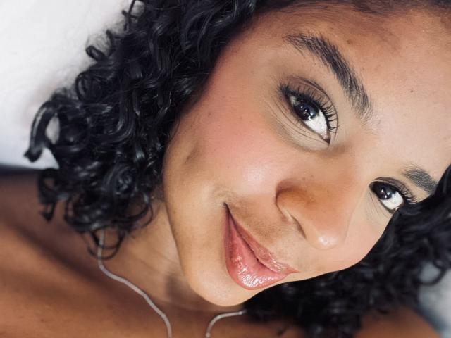 LisaMendez's face, showing curly black hair, brown skin, and a gentle smile with glossy pink lips. Her eyes are slightly upturned, and she's wearing a thin silver necklace.