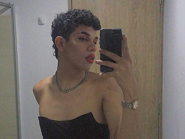 Stefany Colliva, met kort krullend zwart haar, rode lippenstift, een zwarte top zonder bandjes, een ketting, die een foto van zichzelf in de spiegel maakt met haar telefoon, kijkt naar de camera, met een ondeugende uitdrukking, en op de achtergrond is een houten deur te zien..
