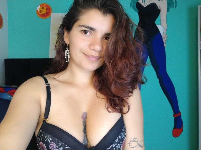 BulmaHot with wavy brown hair, wearing a black floral bra, showing her tits, small tattoo on chest, and a cute smile, wearing earrings, in a teal room.