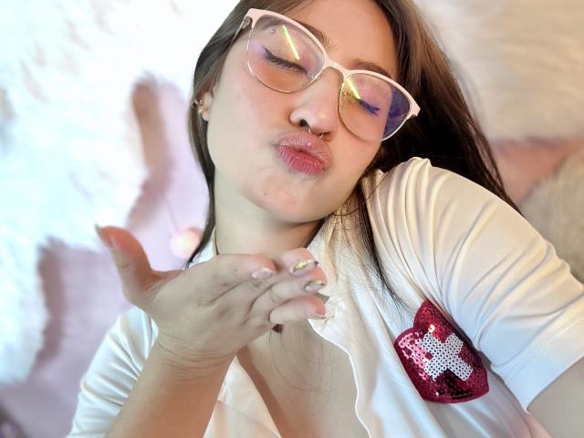LilithAsian with puffy lips, wearing white glasses and a white shirt with a red heart patch, pokes her cheek. Her nails are painted green with a glittery design.