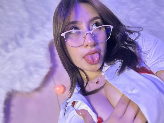 LilithAsian with glasses, tongue out, holding her tits, wearing a white shirt, red bra, and a necklace, with a nose ring, in a purple-lit selfie.