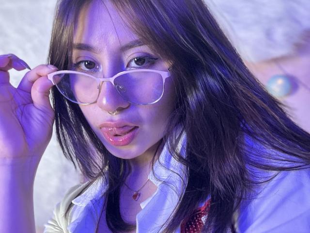 LilithAsian with glossy pink lips, holding white glasses, nose ring, long brown hair, wearing white shirt, blueish-purple light, looking up with slightly parted pink lips, soft expression.