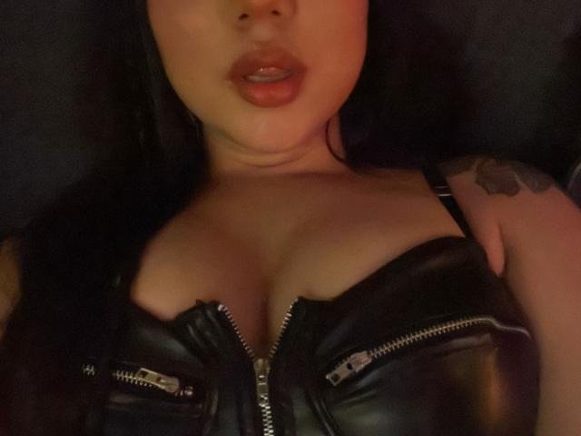 SteffanyTaylor with pouty lips, black leather zipped top, showing big boobs, tattoo on right shoulder, dark hair, slightly blurry, warm lighting, soft skin, dark background.