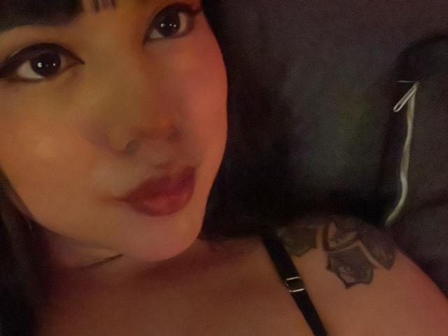 SteffanyTaylor's face with dark lipstick, large eyes, and a tattoo on her right shoulder, wearing a black strap, in dim lighting. Boobs partially visible in the bottom frame.