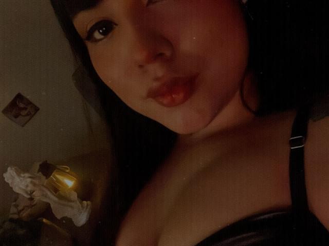 SteffanyTaylor's face with dark hair, slightly parted lips, and a black strap showing her tits. Her skin is warm-toned, and she's in dim light. Her nose is slightly up.