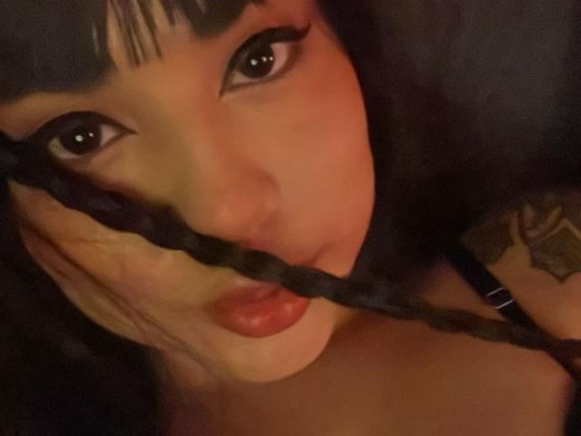 SteffanyTaylor with black braided hair, large brown eyes, and red lips, holding a braid in front of her mouth, showing a tattoo on her right arm.