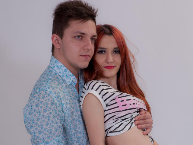 HotCoupleAnal met rood haar, met borsten in een gestreepte shirt, leunt tegen HotCoupleAnal met bruin haar, een blauw geruit shirt, die haar vasthoudt. Beide hebben serieuze, licht sexy uitdrukkingen..