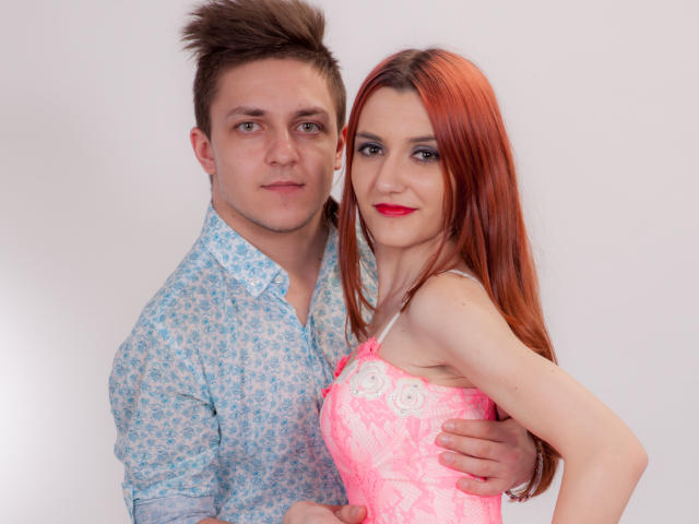 HotCoupleAnal: Hombre con camisa azul estampada con flores, cabello castaño y puntiagudo, sosteniendo a una niña con vestido rosa de encaje, lápiz labial rojo y cabello largo y rojo. La mano de la niña está sobre su trasero..