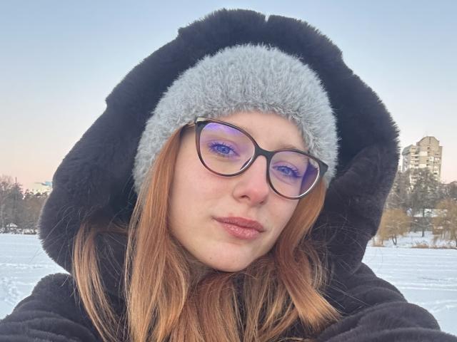 MiOneWallace has a fluffy black hood with gray trim, purple-tinted glasses, and red-brown hair. She's wearing a black fuzzy coat, with a white snowy background.
