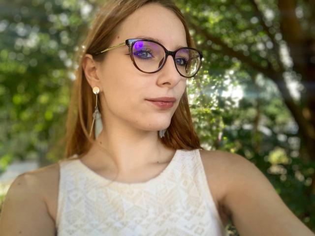 MiOneWallace has light brown hair, wears black glasses, a white lace sleeveless top, and small earrings. She's smiling slightly with a sunlit green background.
