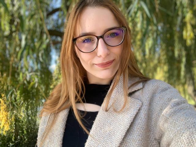 MiOneWallace has pale skin, red hair, and is wearing black-framed glasses with purple lenses. She's in a beige coat, black top, smiling, with green foliage in the background.