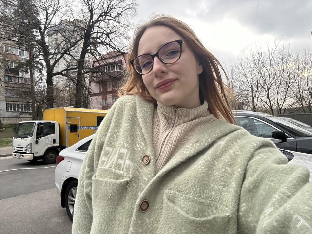 MiOneWallace with glasses, green fuzzy coat, beige turtleneck, and red hair, smiling outside, white car and yellow truck in background, overcast sky, leafless trees.