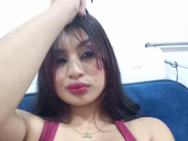 EmmaaRodriguez with dark brown hair, red lipstick, and a gold necklace, holding her head, showing a small bruise under her right eye, sitting on a blue couch.