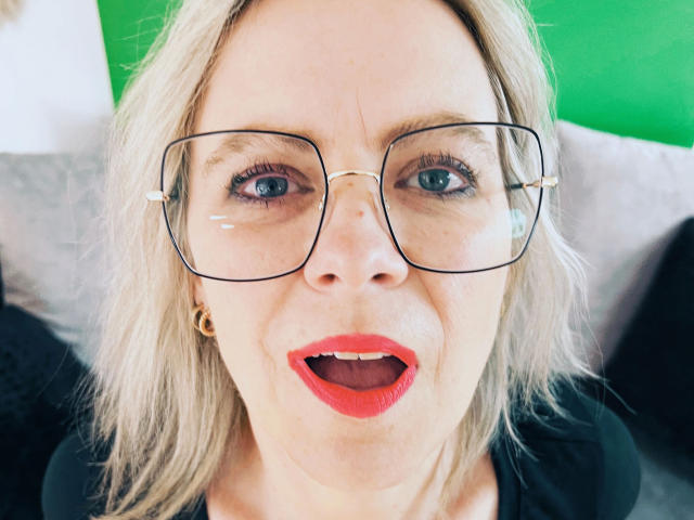 MiaHase-hot with glasses, blonde hair, red lipstick, and gold earrings, mouth open in surprise, blue eyes, black shirt, green background.