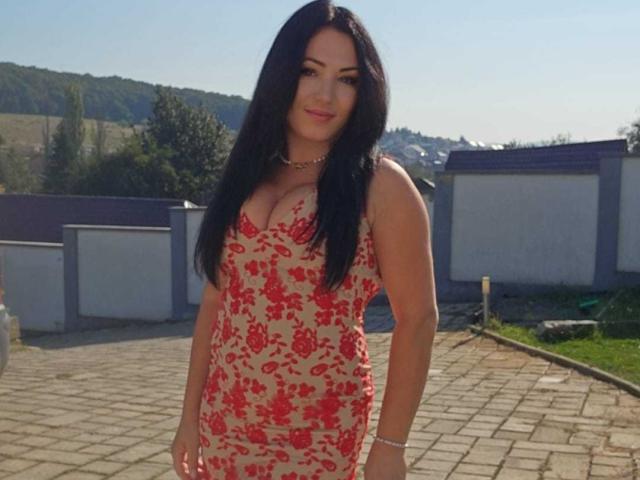 AmelyBrown with long black hair, wearing a red floral dress, showing her tits, standing on a brick patio, smiling, wearing a bracelet, and looking cute.