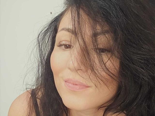 AmelyBrown has messy black hair covering her right eye, brown eyes, and pink lips. She's smiling slightly with a cute, relaxed expression. Her tits are partially visible.