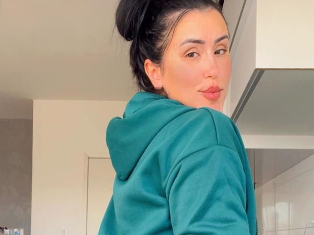 SherllyStar with dark hair in a bun, green hoodie, pouty pink lips, and a slight smile, looking over her shoulder. Her brown eyes are centered in the frame.