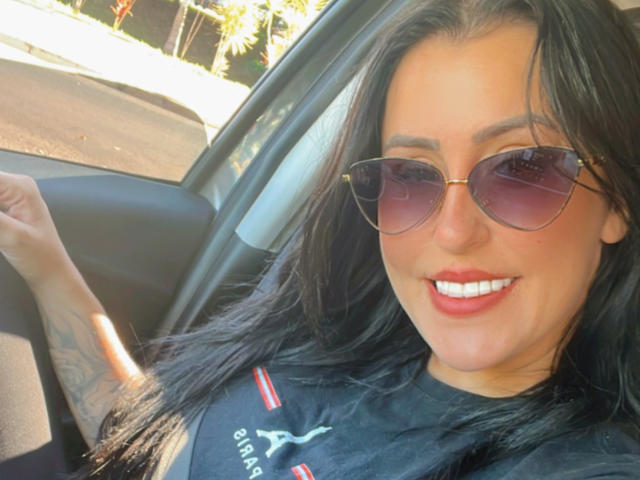 SherllyStar with dark hair, wearing aviator sunglasses, black shirt, and red-white accents, smiling in a car, showing a tattoo on her right arm.
