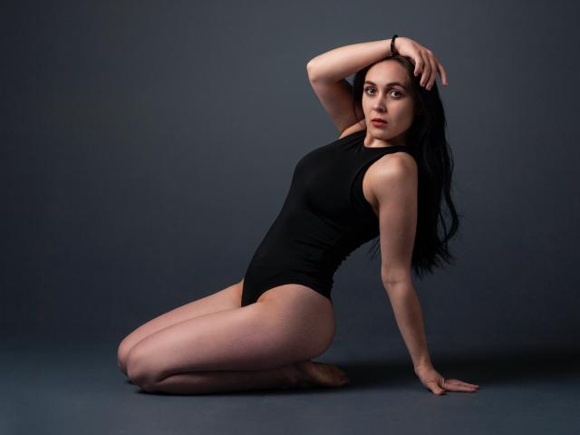 IziPaterson kneels on the floor, wearing a black one-piece. She has pale skin, long black hair, and small tits. Her hand rests on her head, looking up.