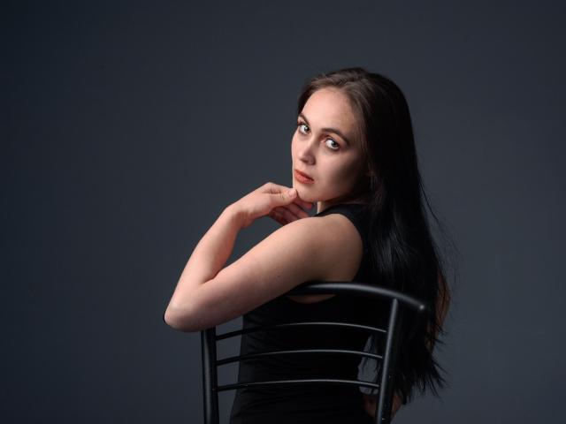 IziPaterson has long black hair, pale skin, and is looking over her shoulder with a slight smile. She's wearing a black sleeveless top, sitting on a black chair.