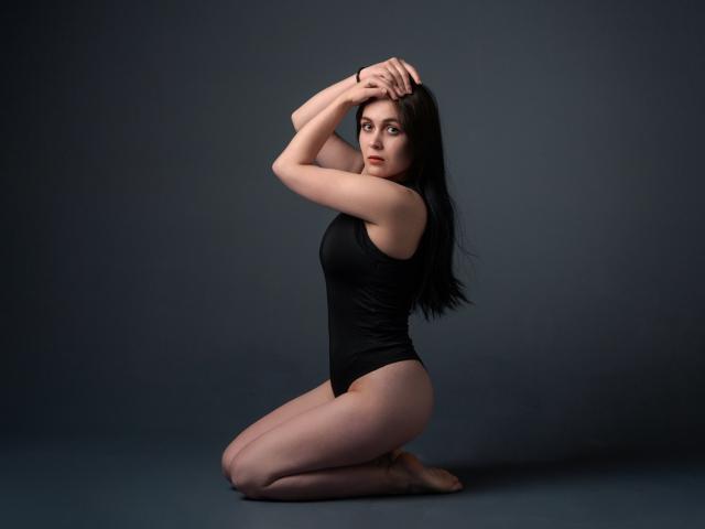 IziPaterson, ajoelhada, vestindo uma camiseta de alça preta, pele clara, cabelo preto longo, braços levantados, olhando para a câmera, mostrando os seios e o bumbum, fundo cinza..