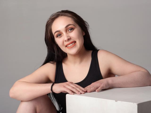 IziPaterson with long brown hair, wearing a black tank top, sitting with arms on a white cube, smiling with orange lipstick, pale skin, and a black wristband.