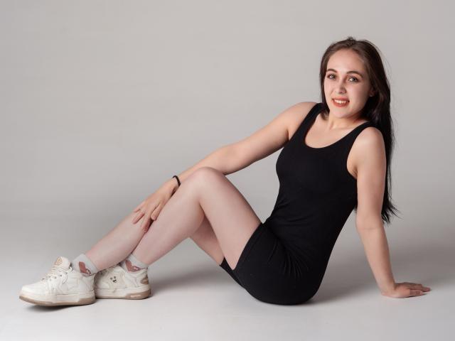 IziPaterson, sitting on floor, black tank dress, white socks, white sneakers, black hair, smiling, arms behind, legs crossed, butt up, hands on floor, gray background.