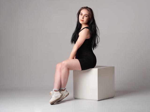IziPaterson with long black hair, pale skin, wearing a tight black dress and white sneakers, sitting on a white cube, legs crossed, looking over her shoulder.