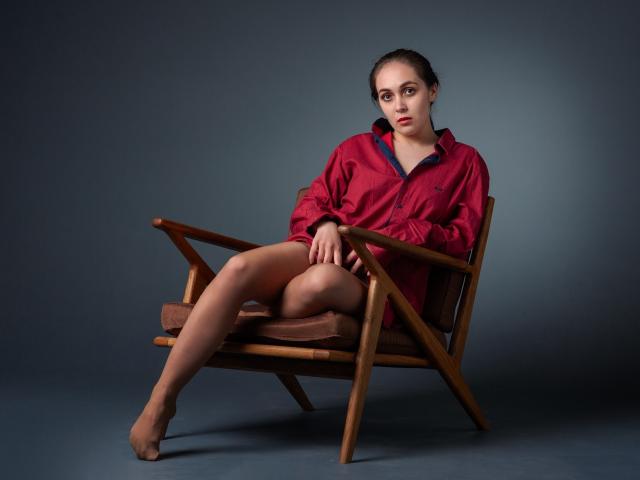 IziPaterson with bare legs, wearing a red jacket, sitting on a wooden chair, legs crossed, arms resting on arms, looking serious, brown hair, no shoes, dark gray background.