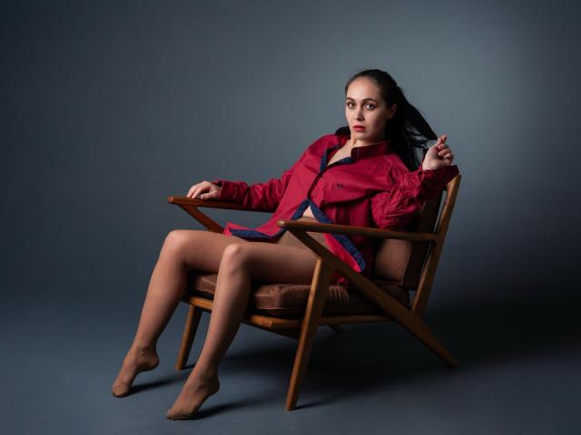 IziPaterson, with brown legs and bare feet, sits in a wooden chair, wearing a red jacket, and holding a cigarette. She has a serious expression and dark hair.
