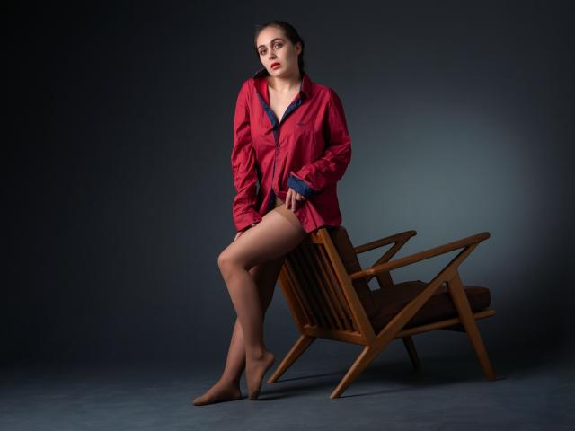 IziPaterson, with brown hair, sits on a wooden chair, wearing a red button-up shirt, no pants, showing her bare legs and feet, looking serious, in a studio.