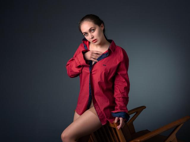 IziPaterson with bare-ass, wearing a red shirt, hand on chest, sitting on a wooden chair, dark gray background, looking at camera, serious expression, pale skin, brown hair.