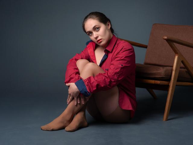 IziPaterson with brown hair, red shirt, no pants, tan socks, sitting on floor, knees up, arms around legs, brown chair behind, serious expression, blue background.