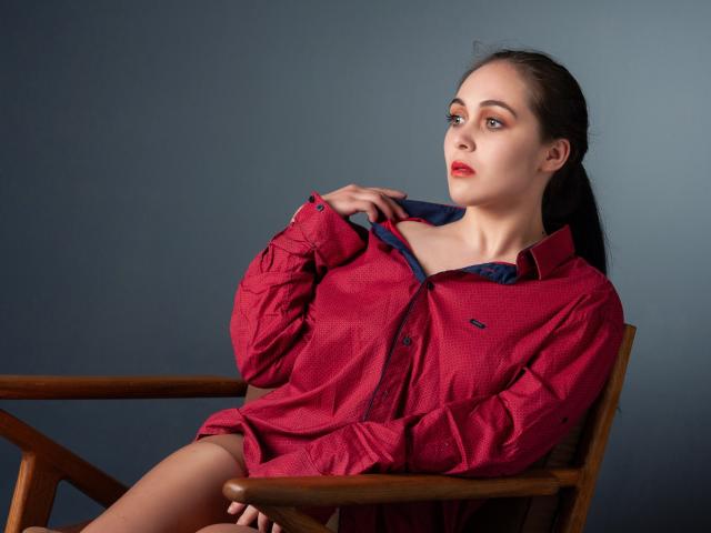 IziPaterson in a red button-up shirt, sitting on a wooden chair, looking to the side with a hand on her collar, showcasing her ass and bare thighs.