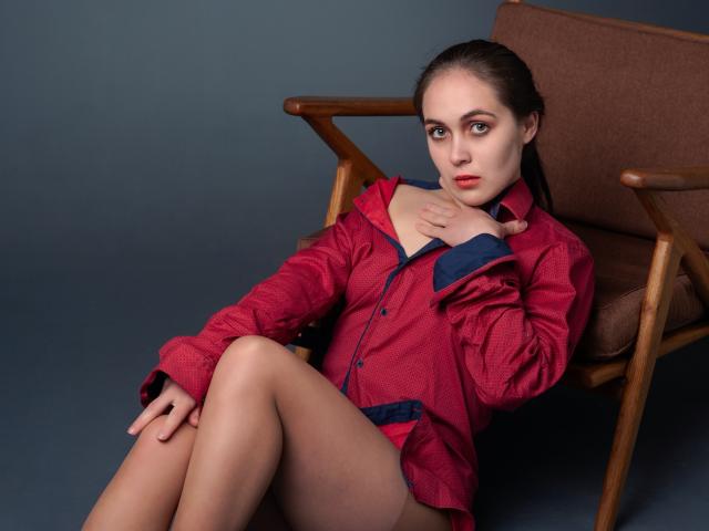 IziPaterson, with red shirt and blue cuffs, sits in brown chair, legs crossed, hand on chest, looking at camera, red lipstick, brown hair, blue-gray background, no pants.