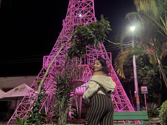 CharloteAdams with dark curly hair, off-shoulder white sweater, and striped pants, standing in front of a pink metal tower at night, showing her curvy ass and looking back.